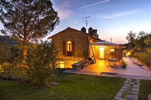 A stone building with a single entrance, a small patio with a table and chairs, and a tree in the foreground. - Agriturismo Antica Quercia Verde - La Fonte (Cortona)