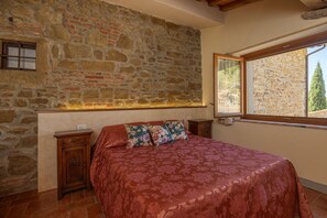 Une chambre avec un mur en pierre, un lit recouvert d’une literie à motifs floraux, une table de chevet et une fenêtre donnant sur un paysage verdoyant.