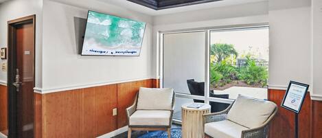A hotel lobby with a television mounted on the wall, a seating area with two chairs and a round table, and a large window offering a view of the outdoors.