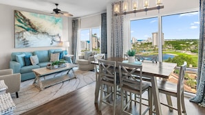 A modern living room with a large window, a dining area, and a ceiling fan. - Luau I 7010/7012 - Sunset Suite (Miramar Beach)