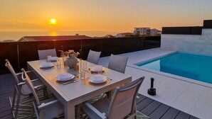 Un área de comedor al aire libre con piscina, mesa y sillas blancas, todo silueteado contra un atardecer.