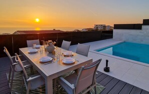 An outdoor dining area with a pool, white table, and chairs, set against a sunset backdrop. - Villa Mandre Hills II (Mandre)