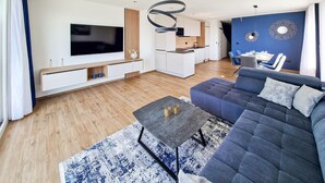Ein modernes Wohnzimmer mit großem Fernseher, einer Couch, einem Couchtisch und einer Küchenzeile.