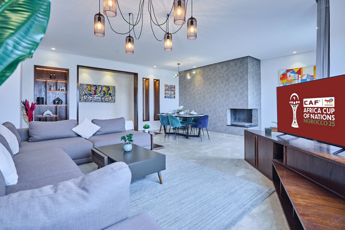 A modern living room with a sofa, coffee table, dining area, and a TV displaying the CAF Africa Cup of Nations logo.