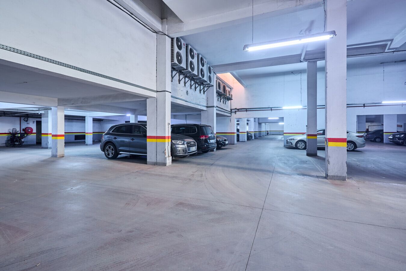 Um estacionamento multi andar com pilares de concreto, iluminação fluorescente e vários carros estacionados.