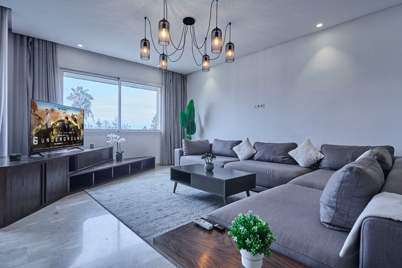 A modern living room with a large window, grey sofa, coffee table, and a TV displaying a movie poster.