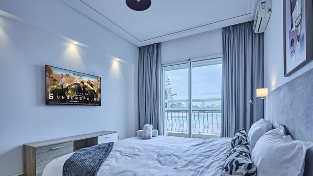 Ett hotellrum med en stor säng, ett nattduksbord, en tv som är monterad på väggen och ett fönster med gardiner.