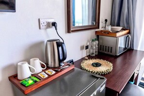 A hotel room with a kitchenette, a coffee maker, a kettle, a microwave, a small refrigerator, a tray with coffee cups and tea bags, a woven placemat, and a mirror above the counter. - Hammock Suites MUP - by Zenbreak (Castries)