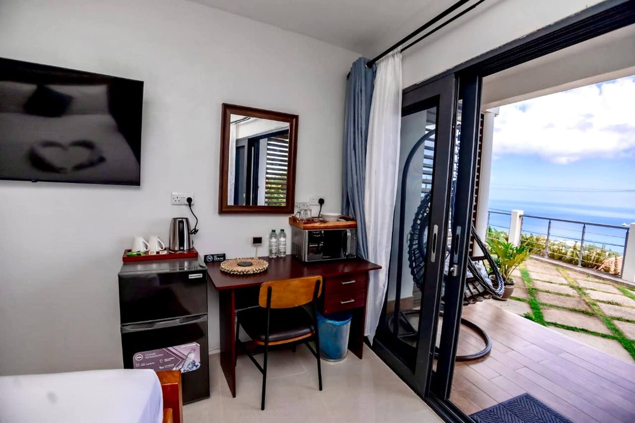 Ein Hotelzimmer mit Bett, Schreibtisch mit Stuhl, Kühlschrank, Kaffeemaschine, Spiegel und Meerblick durch eine Glastür.