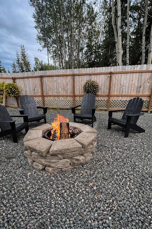 A stone fire pit with four black Adirondack chairs around it, set on gravel with a wooden fence and trees in the background.