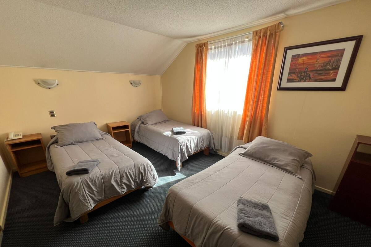 Habitación de hotel con tres camas individuales, una mesita, una silla, un cuadro en la pared y una ventana con cortinas.