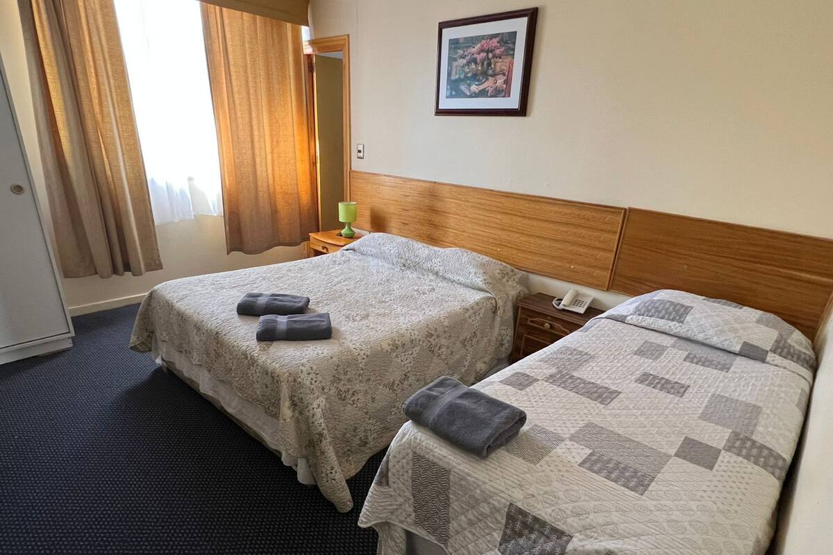 Habitación de hotel con dos camas, una mesita de noche con lámpara, un cuadro enmarcado en la pared y una ventana con cortinas.