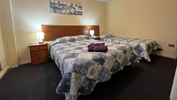 Habitación de hotel con dos camas, cabecero, mesita de noche, lámpara y un cuadro en la pared.