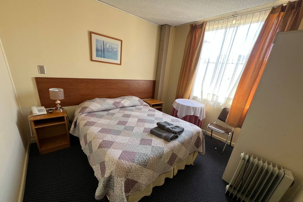 Habitación de hotel con cama, mesita de noche con lámpara, silla, una mesita pequeña, radiador y ventana con cortinas.