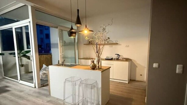 A modern kitchen with a central island, clear bar stools, pendant lighting, and a sliding glass door leading to a balcony.