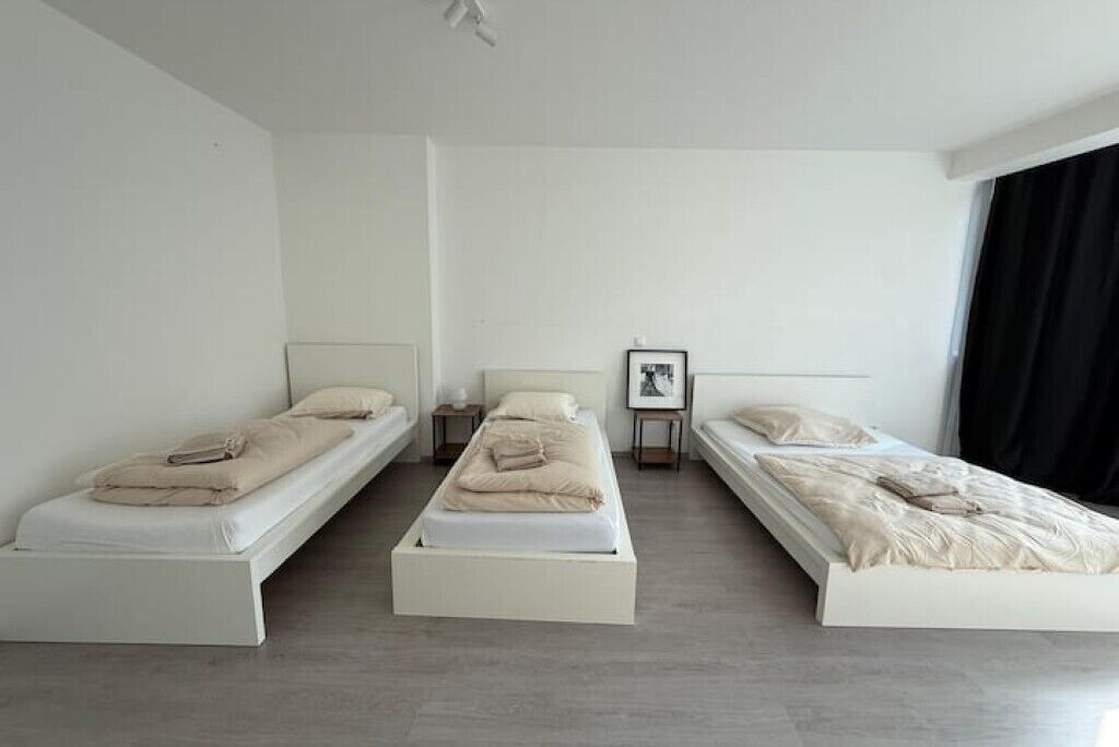 Quarto de hotel minimalista com três camas de solteiro, uma pequena mesa de cabeceira com luminária e um quadro na parede.