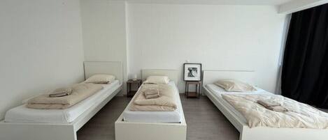 Quarto de hotel minimalista com três camas de solteiro, uma pequena mesa de cabeceira com luminária e um quadro na parede.