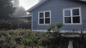 Una casa con fachada azul, dos ventanas y un jardín con diversas plantas y un bebedero para pájaros.