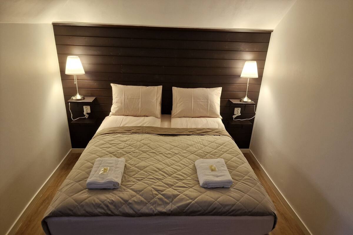 A neatly made bed with a quilted headboard, two bedside lamps, and two folded towels on the footboard.