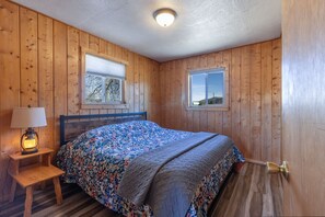 Ein Zimmer mit Holzwänden, einem Bett, einem Nachttisch, einer Lampe und zwei Fenstern.