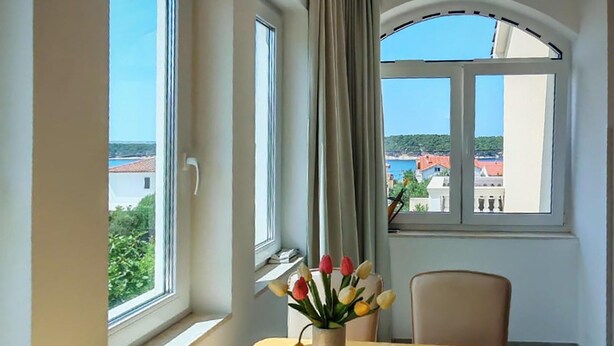 A room with a table, a chair, a vase with flowers, and a view of a coastal town through large windows. - 1 bedroom nice apartment in Barbat (Barbat)