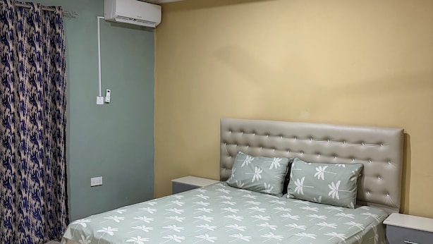 Un dormitorio con cama, ventilador de techo, unidad de aire acondicionado, mesita de noche y velador.