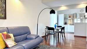 Ein modernes Wohnzimmer mit Sofa, Esstisch mit Stühlen, Küchenbereich und einer großen Stehlampe.