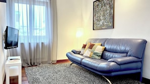 A living room with a blue leather sofa, a flat-screen TV, a white side table, a patterned rug, and a window with sheer curtains. - Entire Apartment for 5 with parking near Syracuse Greek Theater (Siracusa)