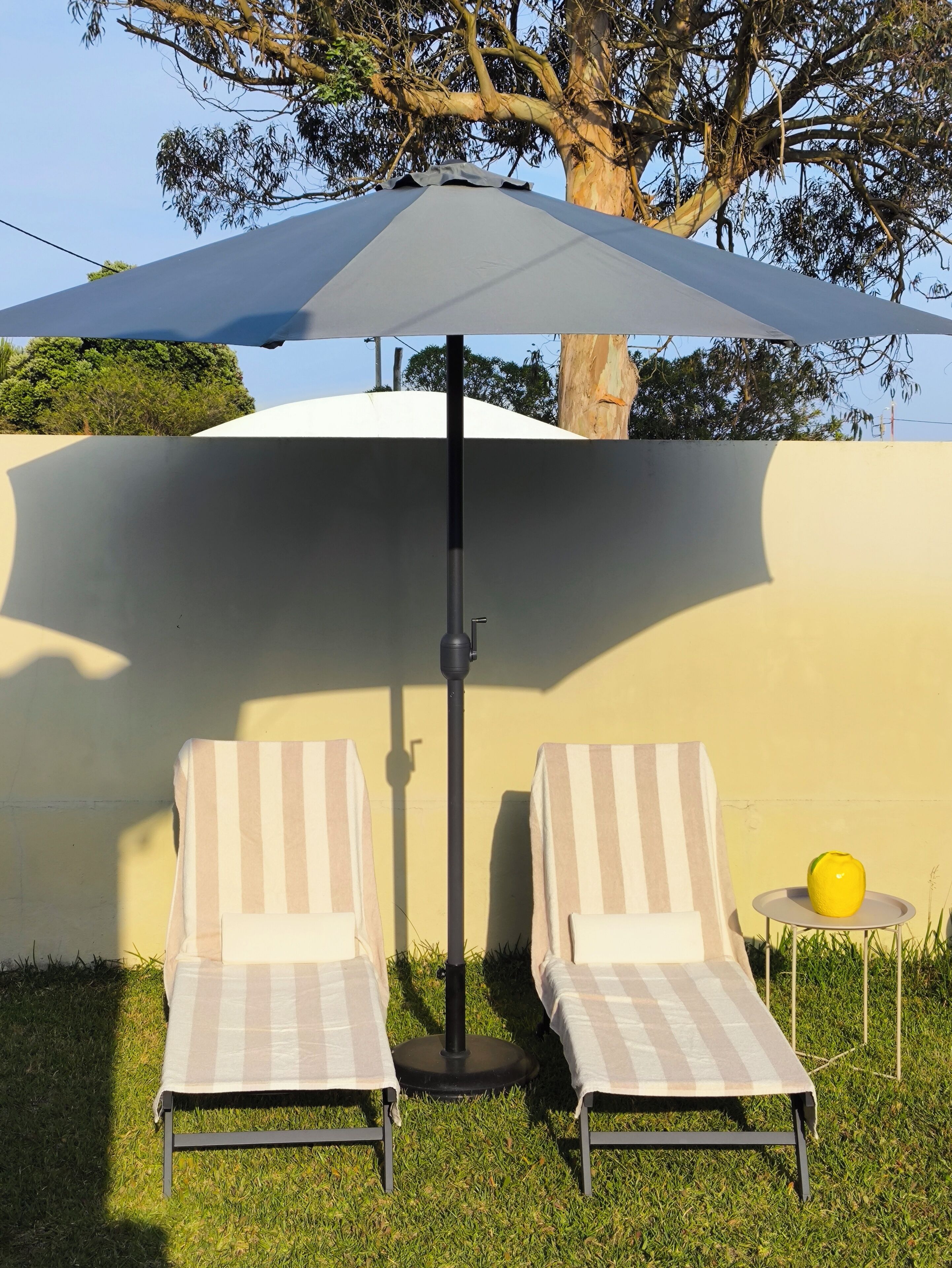 Two lounge chairs with striped cushions, a round table with a yellow object, and a large umbrella providing shade.
