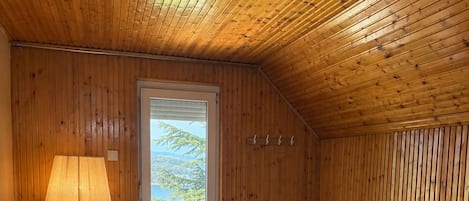 A bedroom with a wooden ceiling, a bed with white linens, a lamp, and a view through a door.