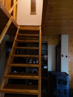 Una escalera de madera que lleva a un altillo con una cama de altillo, un estante pequeño con adornos y un calentador negro.