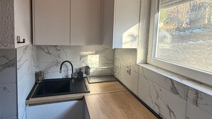 Une cuisine compacte avec des meubles blancs, un plan de travail en bois, un évier et une fenêtre donnant sur les arbres.
