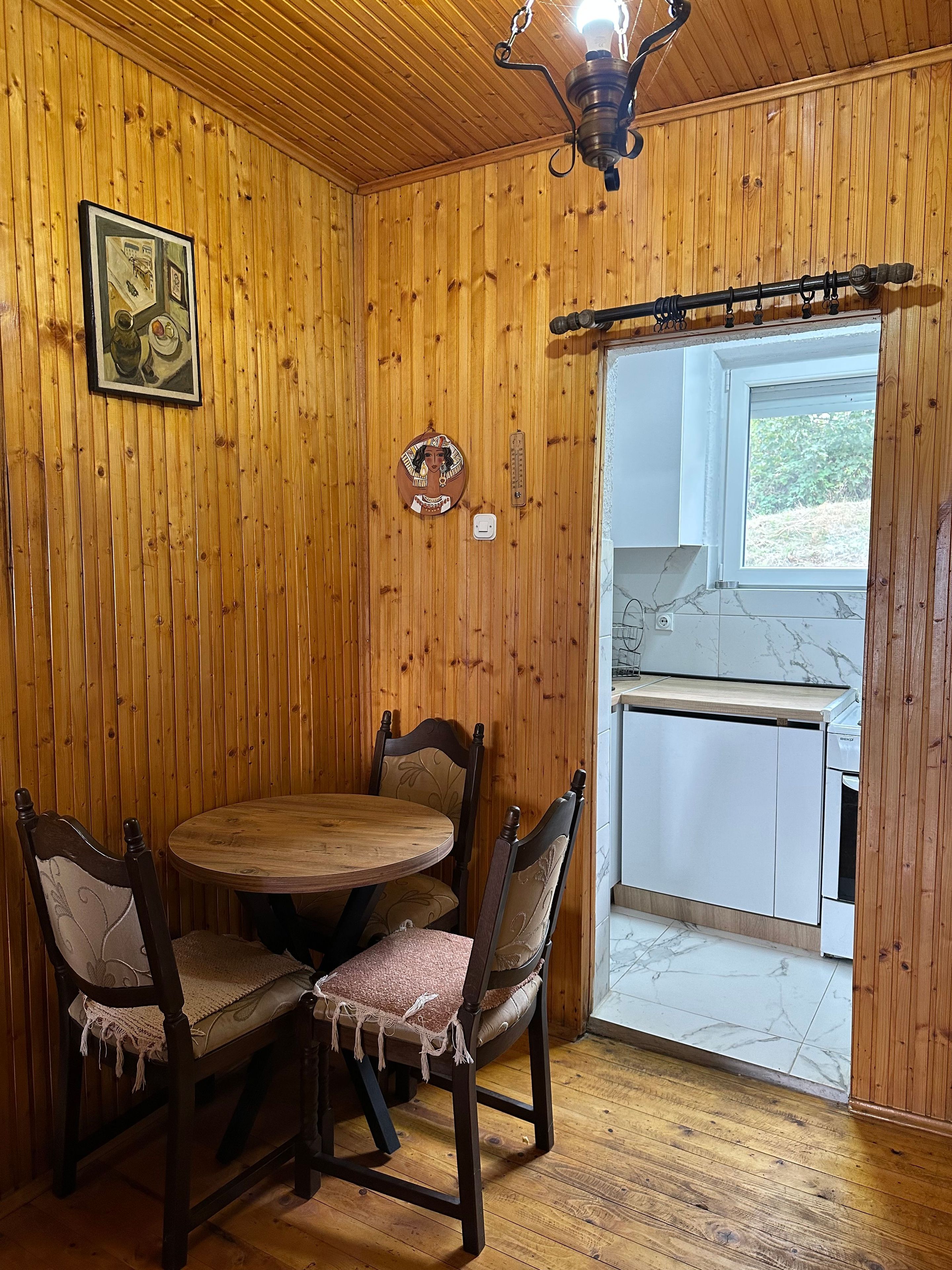 A wooden-paneled room with a dining table and chairs, a light fixture, and a window leading to a kitchen area.