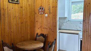 A wooden-paneled room with a dining table and chairs, a light fixture, and a window leading to a kitchen area.