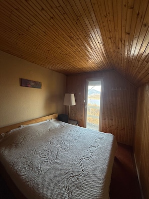 A wooden-paneled room with a bed, a nightstand, a lamp, and a view of the outdoors. - Mavrovo Lake View Chalet (Leunovo)