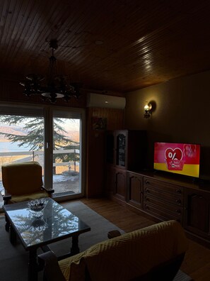 Una sala de estar con una mesa de vidrio, un sofá, un televisor mostrando una oferta y una vista de un paisaje montañoso a través de un puerta corrediza de vidrio.