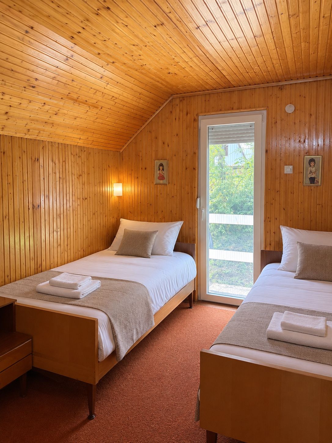 A room with two beds, wooden walls, a door leading outside, and a small wall-mounted light fixture.