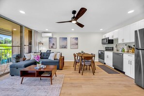 Eine moderne Küche mit weißen Schränken, ein Essbereich mit Holztisch und -stühlen, eine Couch und ein Deckenventilator.