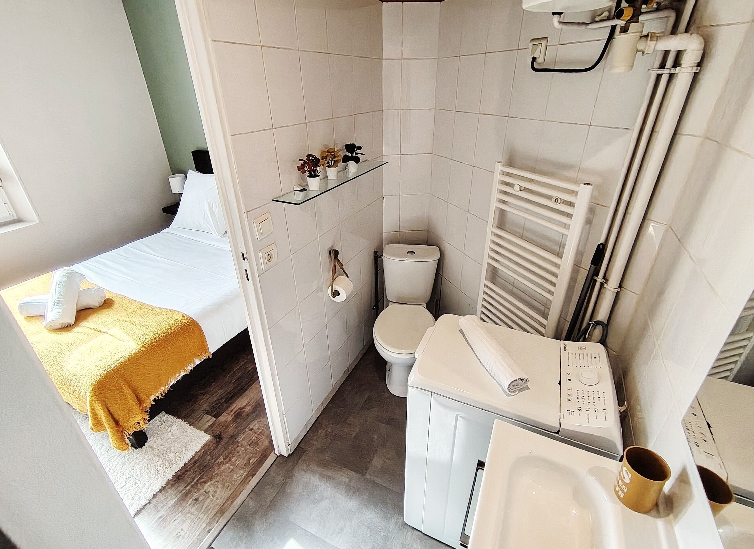 A compact hotel room with a bed, a small bedside table, a toilet, a sink, and a shower area.