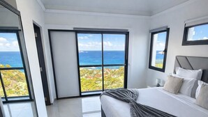 A modern bedroom with a large bed, a view of the ocean, and a sliding glass door leading to a balcony.