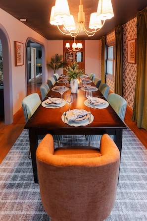 A dining room with a long table set for six, surrounded by chairs, with a chandelier above and a plant centerpiece.