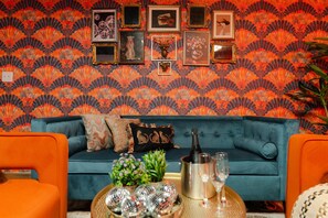 A teal sofa with decorative pillows, a small table with a bottle and glasses, and a wall with framed pictures.