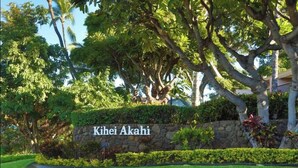 Eine Steinmauer mit einem Schild, das 'Kihei Akahi' zeigt, umgeben von üppigem Grün und Bäumen.