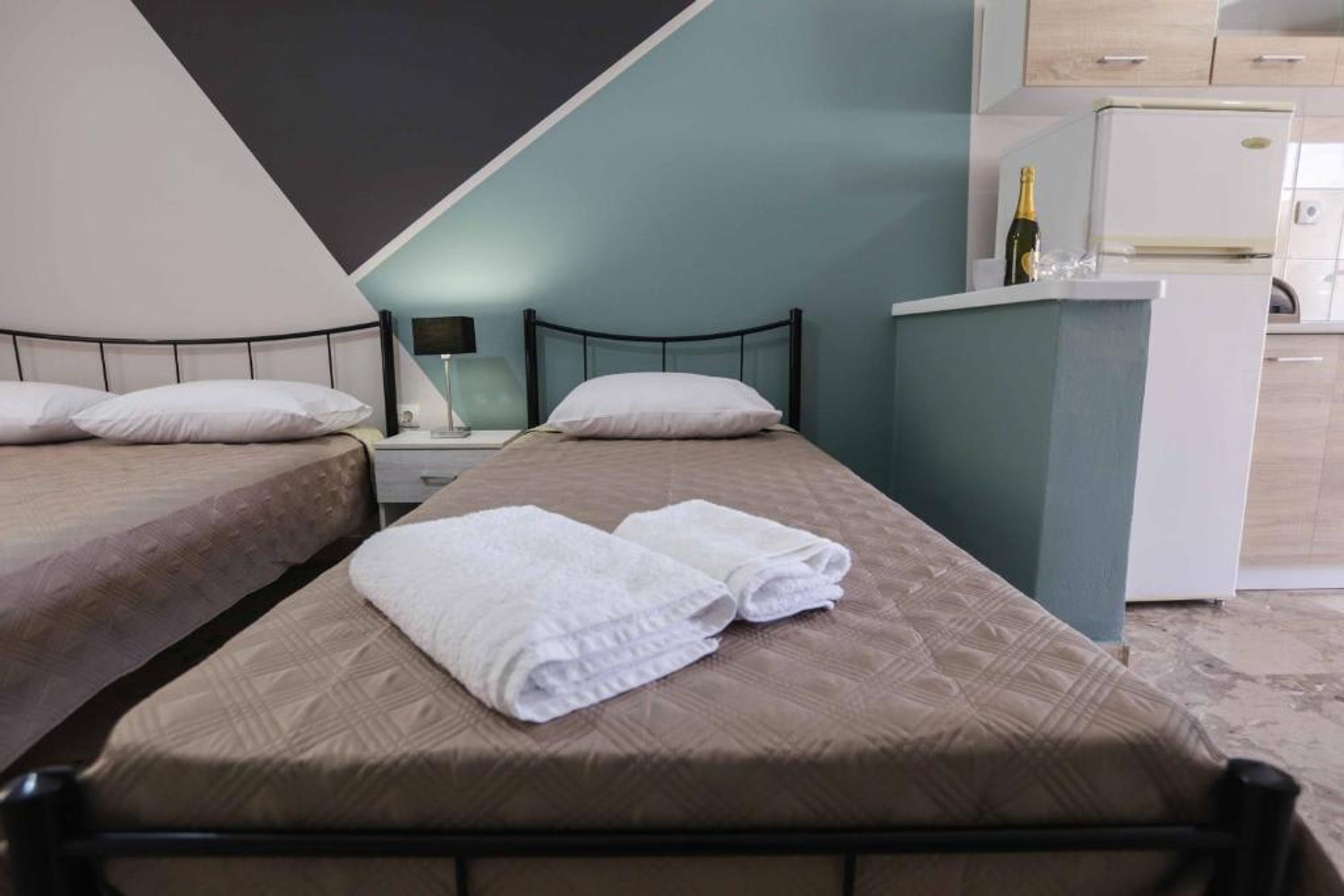 A hotel room with two beds, a minibar, a lamp, and a bottle of champagne.
