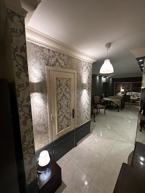 Une pièce avec un papier peint à motifs, une porte, un coin repas avec des chaises et une table, et un luminaire suspendu.