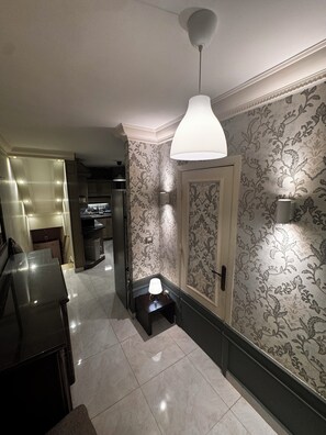Un couloir avec un papier peint à motifs, une porte, une petite table avec une lampe et un luminaire au plafond.