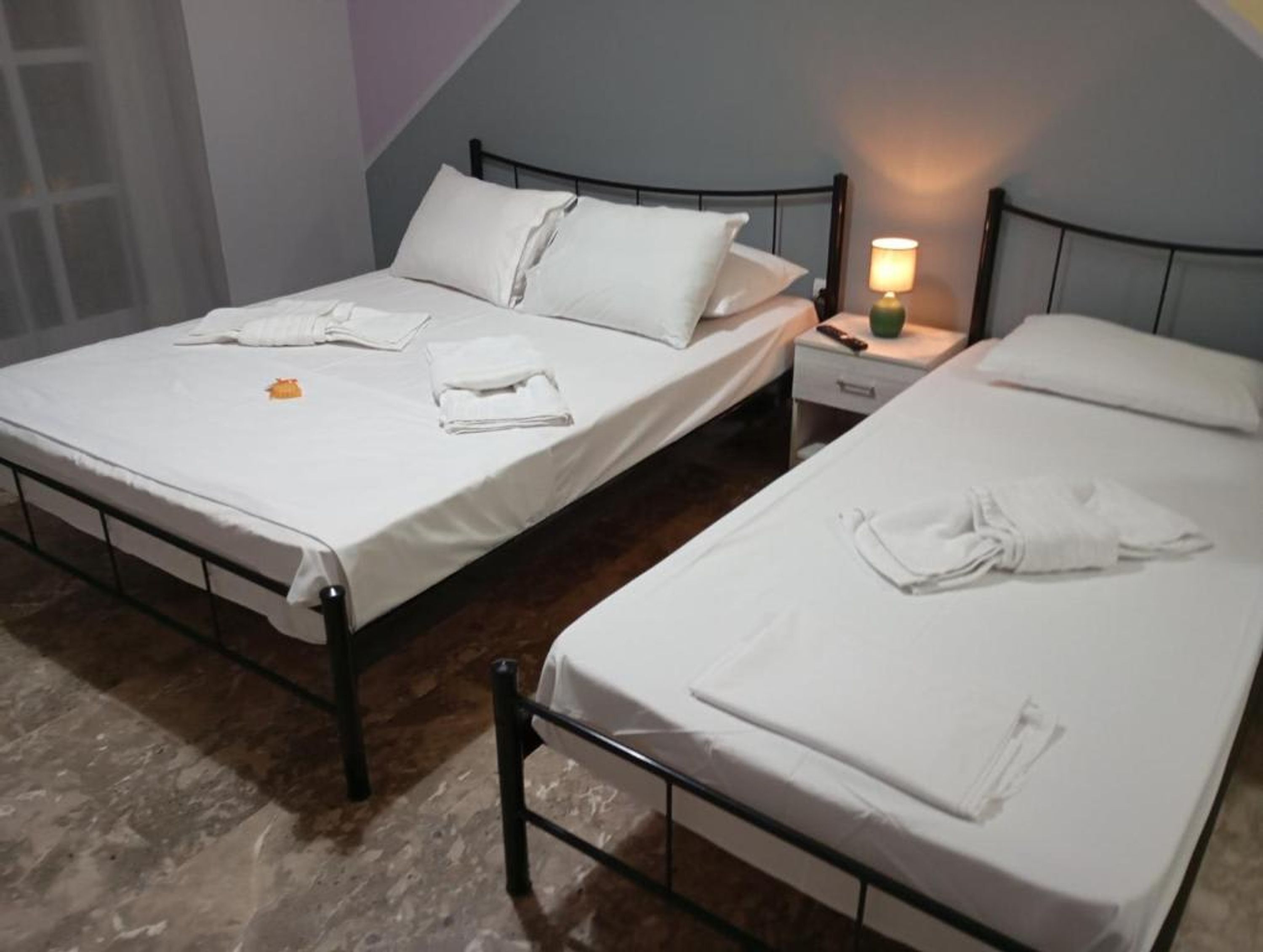 Two single beds with white linens, a nightstand with a lamp, and a window with a view of the outside.