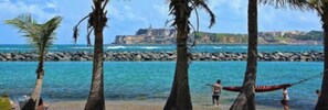 A beach with palm trees, a rocky breakwater, and a distant building. - Villa De Gloria II - Elevated Signature Stay (Bayamon)