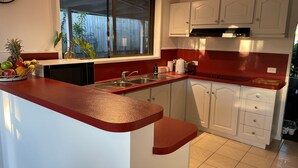 Een keuken met witte kasten, een rode aanrecht en een raam met uitzicht op een houten schutting, plus een fruitschaal op het aanrecht.