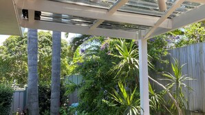 Een overdekt buitengebied met houten omheining, verschillende planten en een constructie met metalen dak.
